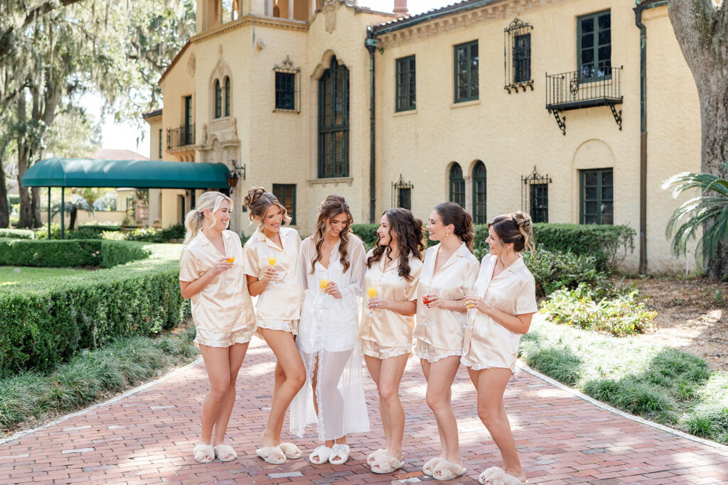 bride wearing white getting ready pajamas and bridesmaids wearing peach pajamas holding mimosas outside of Epping Forest Yacht Club wedding venue in Jacksonville, Florida, captured by Snapshots by Gabriela