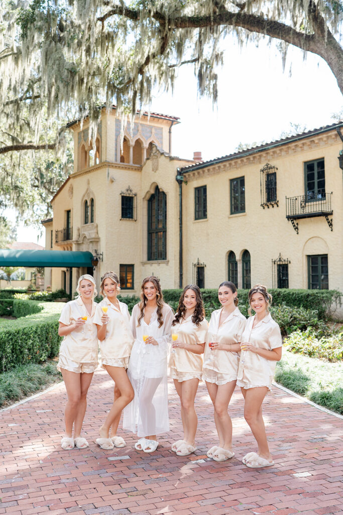bride wearing white getting ready pajamas and bridesmaids wearing peach pajamas holding mimosas outside of Epping Forest Yacht Club wedding venue in Jacksonville, Florida, captured by Snapshots by Gabriela