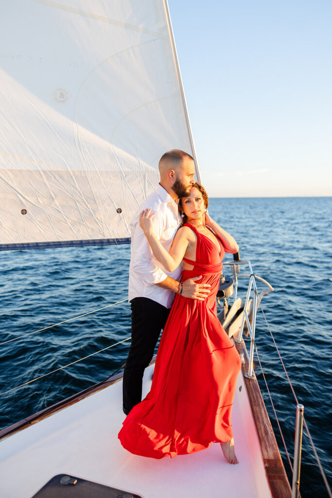 A woman in a red flowing dress with curly brown hair and a man in white romantically embrace on the deck overlooking the water during their sailboat engagement photos, captured by Tampa engagement photographer Snapshots by Gabriela