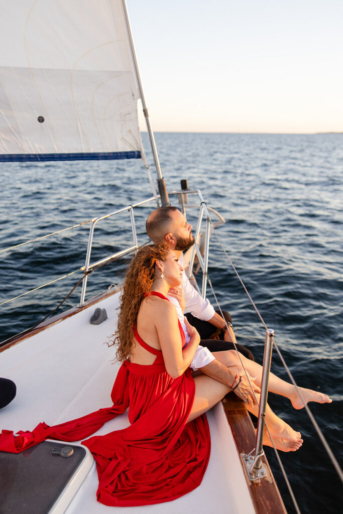 A woman in a red flowing dress with curly brown hair and a man in white romantically embrace on the deck of a sailboat during their sailboat engagement photos, captured by Tampa engagement photographer Snapshots by Gabriela