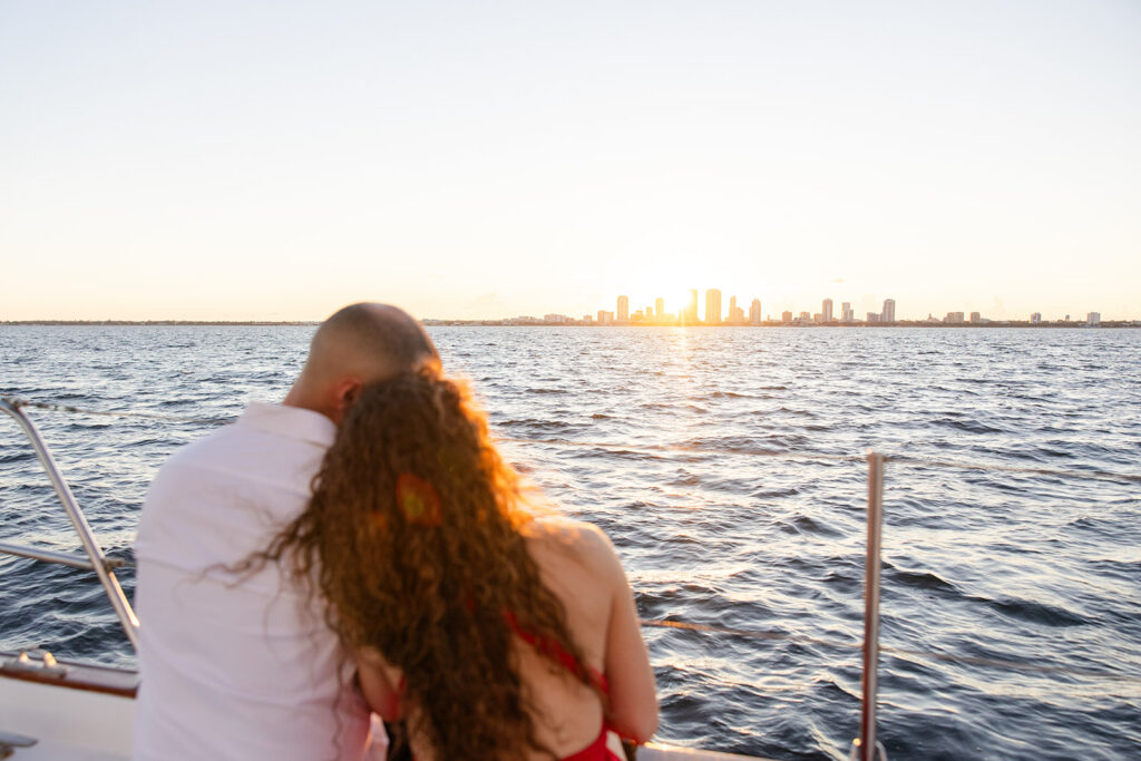 A woman in a red flowing dress with curly brown hair and a man in white romantically embrace on the deck overlooking the water during their sailboat engagement photos, captured by Tampa engagement photographer Snapshots by Gabriela