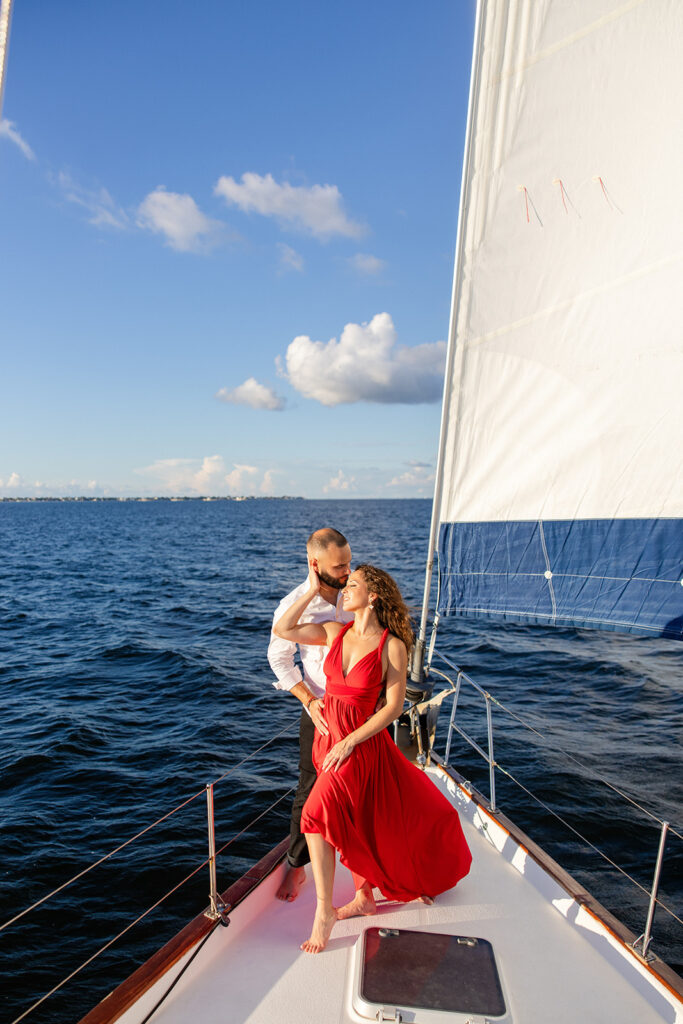 A woman in a red flowing dress with curly brown hair and a man in white romantically embrace on the deck of a sailboat during their sailboat engagement photos, captured by Tampa engagement photographer Snapshots by Gabriela