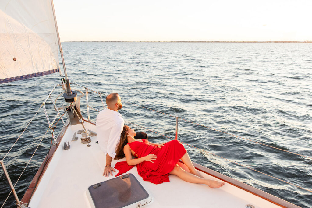 A woman in a red flowing dress with curly brown hair and a man in white romantically embrace on the deck overlooking the water during their sailboat engagement photos, captured by Tampa engagement photographer Snapshots by Gabriela