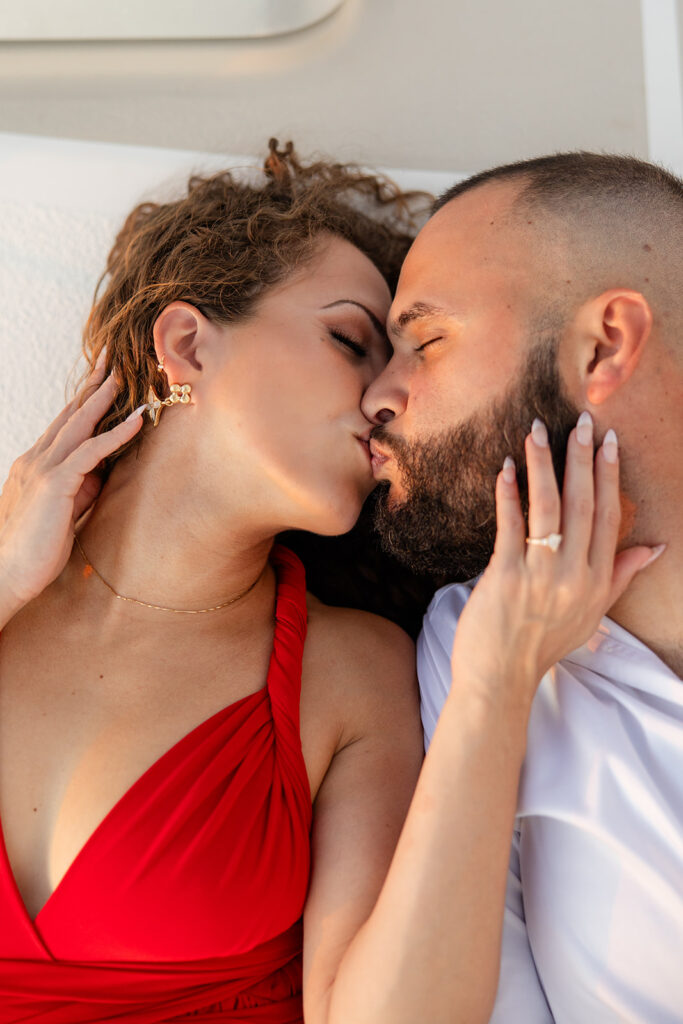 A woman in a red flowing dress with curly brown hair and a man in white romantically kiss with hands on each other's face during their sailboat engagement photos, captured by Tampa engagement photographer Snapshots by Gabriela