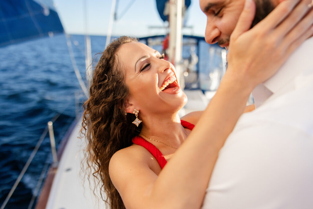 A woman in a red flowing dress with curly brown hair and a man in white playfully embrace on the deck during their sailboat engagement photos, captured by Tampa engagement photographer Snapshots by Gabriela