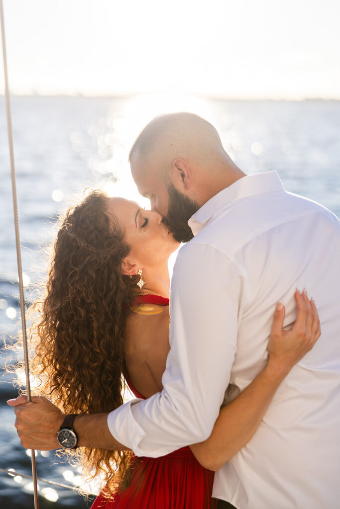 A woman in a red flowing dress with curly brown hair and a man in white romantically embrace in front of the glowing sun during their sailboat engagement photos, captured by Tampa engagement photographer Snapshots by Gabriela
