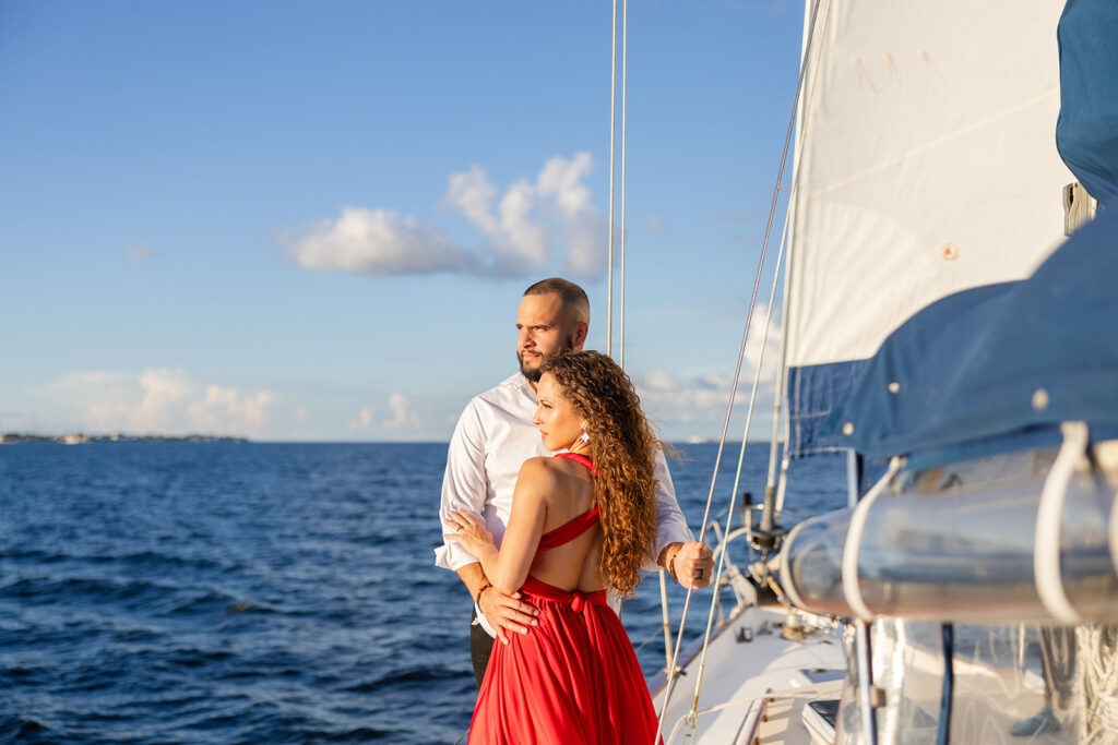 A woman in a red flowing dress with curly brown hair and a man in white romantically embrace on the deck overlooking the water during their sailboat engagement photos, captured by Tampa engagement photographer Snapshots by Gabriela