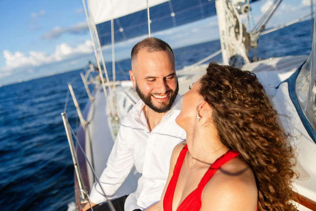 A woman in a red flowing dress with curly brown hair and a man in white give a playful laugh on the deck overlooking the water during their sailboat engagement photos, captured by Tampa engagement photographer Snapshots by Gabriela