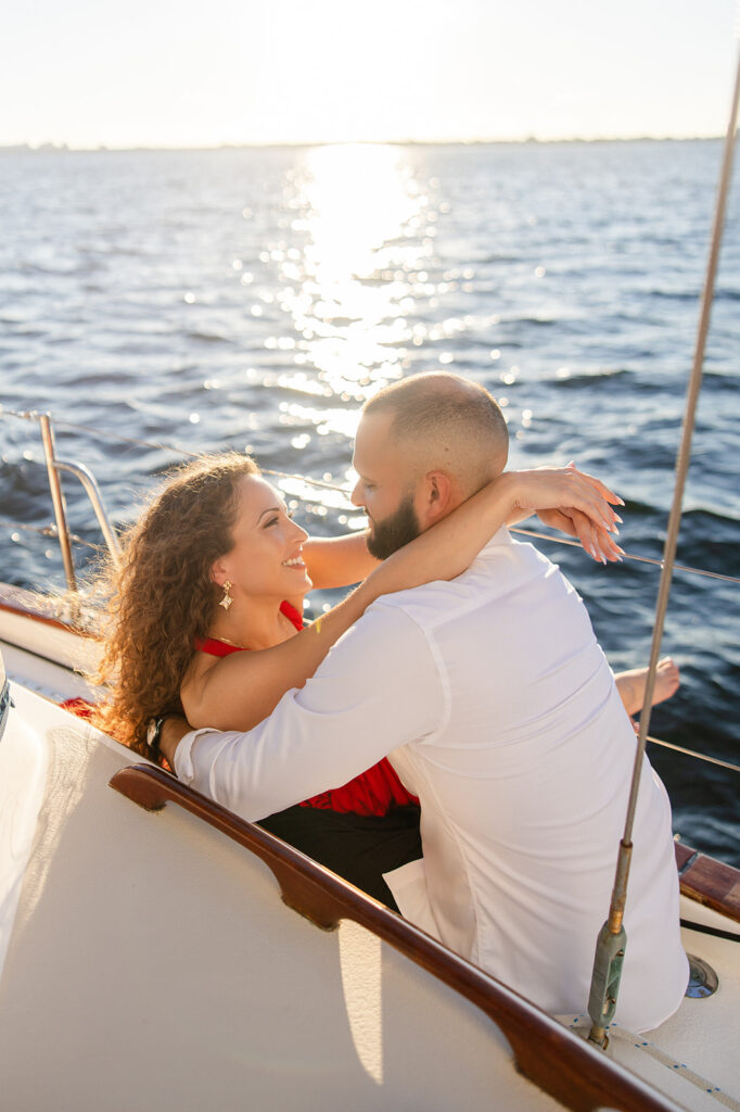 A woman in a red flowing dress with curly brown hair and a man in white romantically embrace in front of the glowing sun during their sailboat engagement photos, captured by Tampa engagement photographer Snapshots by Gabriela