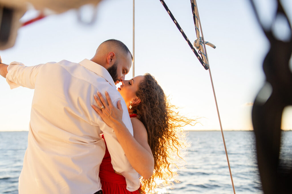 A woman in a red flowing dress with curly brown hair and a man in white romantically embrace in front of the glowing sun during their sailboat engagement photos, captured by Tampa engagement photographer Snapshots by Gabriela