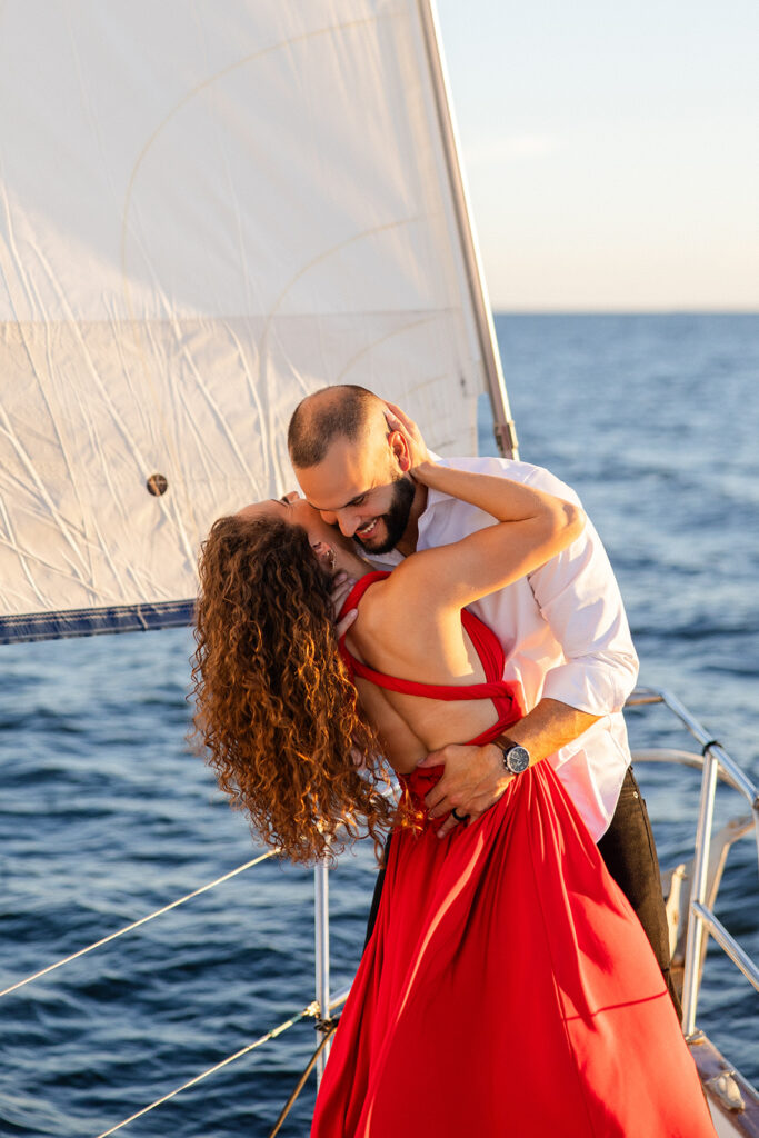 A woman in a red flowing dress with curly brown hair and a man in white romantically embrace on the deck overlooking the water during their sailboat engagement photos, captured by Tampa engagement photographer Snapshots by Gabriela