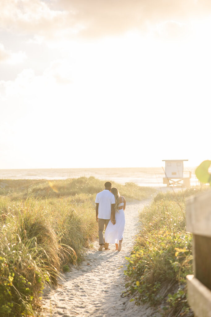 couple wearing white walk along a sandy path in a beach engagement photo at Jetty Park Beach in Cape Canaveral, Florida, captured by Snapshots by Gabriela