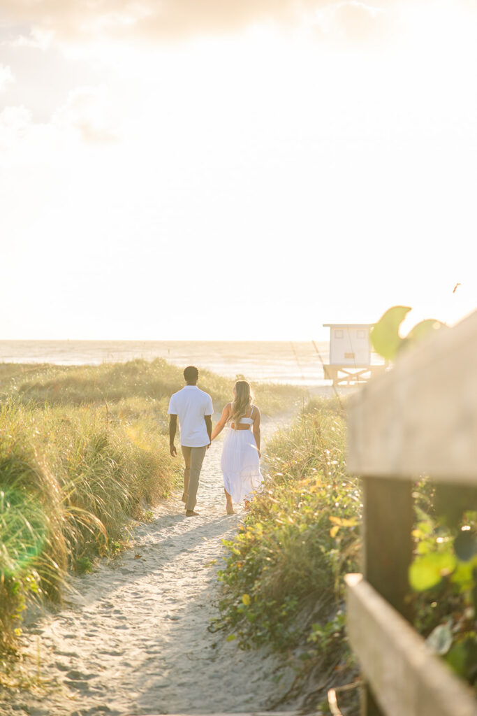 couple wearing white walk along a sandy path in a beach engagement photo at Jetty Park Beach in Cape Canaveral, Florida, captured by Snapshots by Gabriela