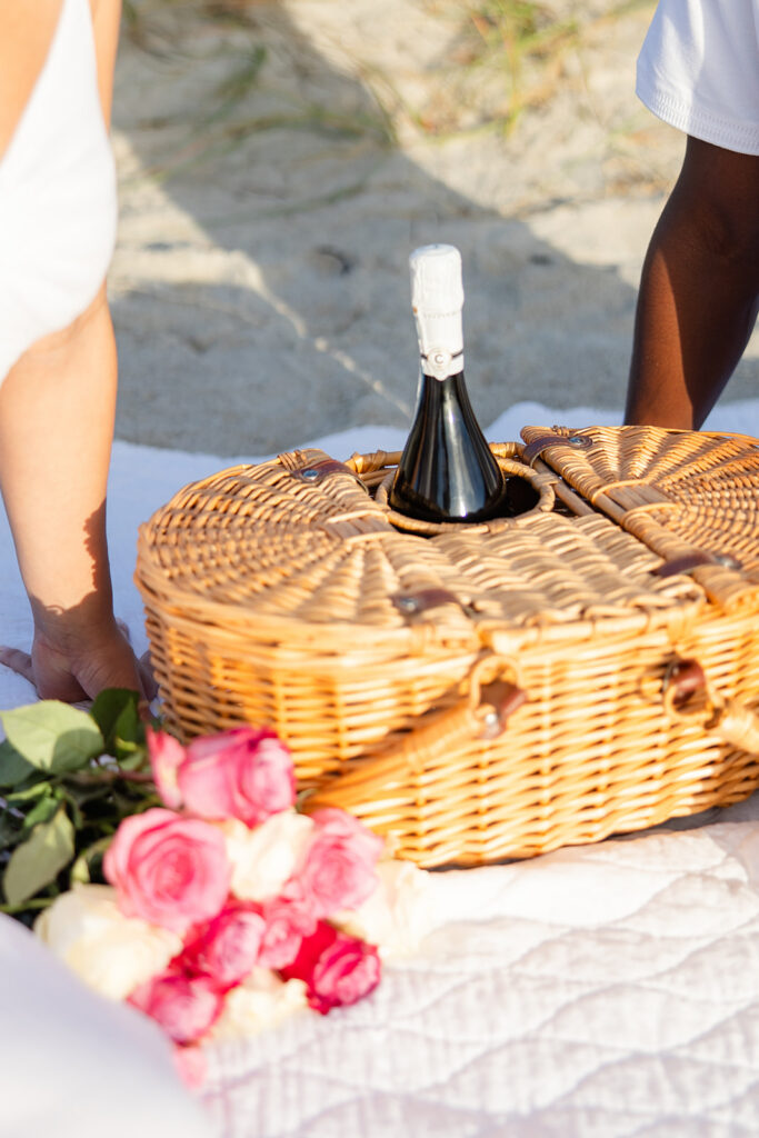 picnic basket with pink and white roses and a champagne bottle on a white blanket in a beach engagement photo at Jetty Park Beach in Cape Canaveral, Florida, captured by Snapshots by Gabriela