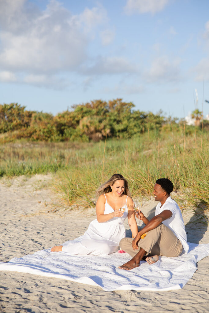 couple clink champagne glasses on a picnic blanket in the sand in a beach engagement photo at Jetty Park Beach in Cape Canaveral, Florida, captured by Snapshots by Gabriela