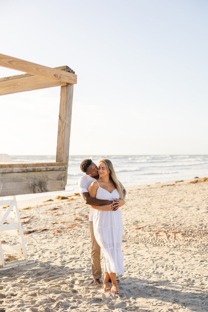couple wearing white embrace in a beach engagement photo at Jetty Park Beach in Cape Canaveral, Florida, captured by Snapshots by Gabriela