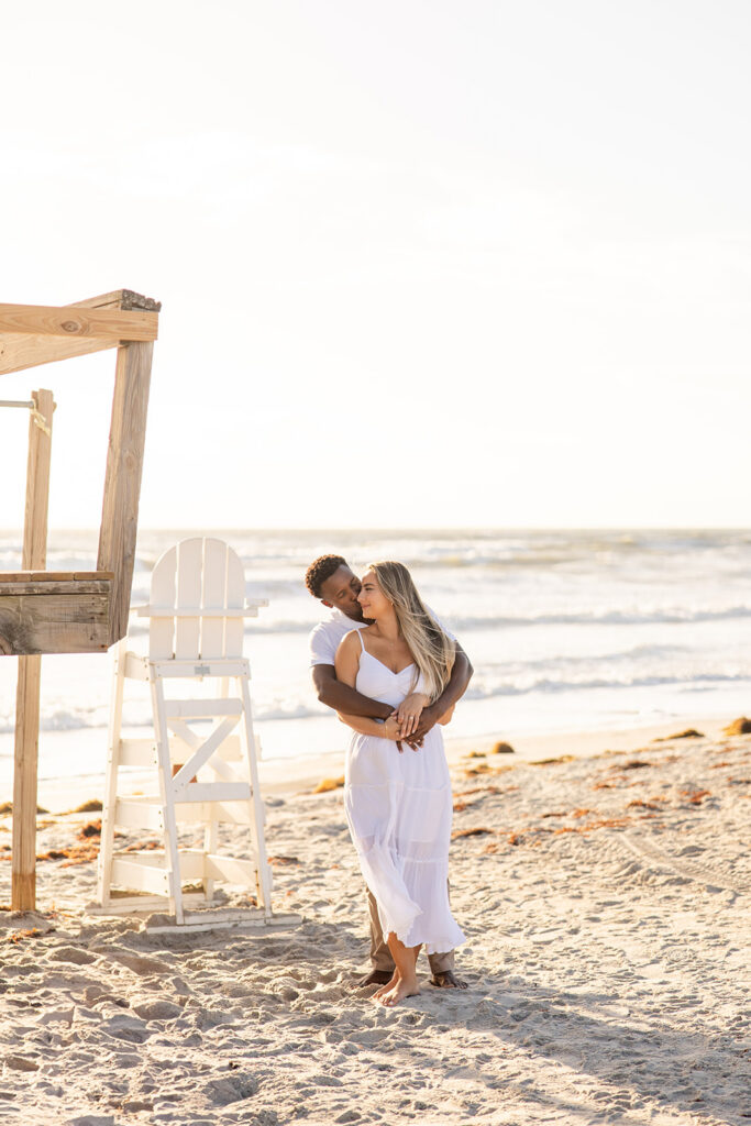 couple wearing white embrace in a beach engagement photo at Jetty Park Beach in Cape Canaveral, Florida, captured by Snapshots by Gabriela