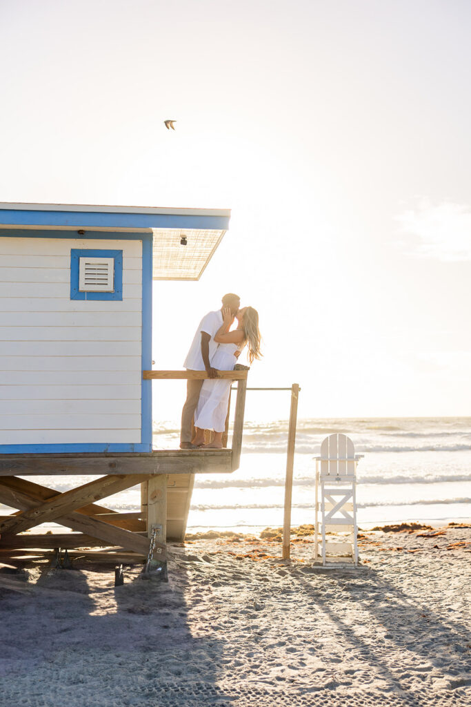 couple wearing white kiss on a lifeguard stand in a beach engagement photo at Jetty Park Beach in Cape Canaveral, Florida, captured by Snapshots by Gabriela