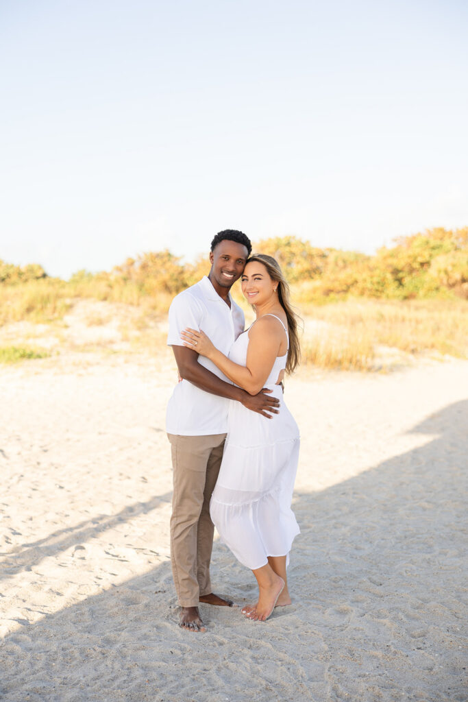 couple wearing white embrace in a beach engagement photo at Jetty Park Beach in Cape Canaveral, Florida, captured by Snapshots by Gabriela