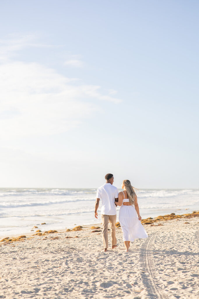 couple wearing white walk along the shore in a beach engagement photo at Jetty Park Beach in Cape Canaveral, Florida, captured by Snapshots by Gabriela