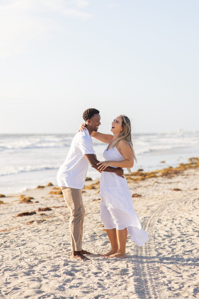 couple wearing white laugh together in a beach engagement photo at Jetty Park Beach in Cape Canaveral, Florida, captured by Snapshots by Gabriela