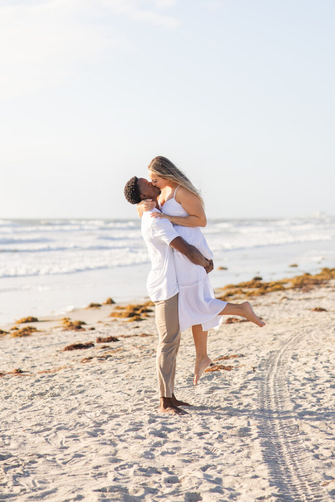 couple wearing white kiss along the shore in a beach engagement photo at Jetty Park Beach in Cape Canaveral, Florida, captured by Snapshots by Gabriela