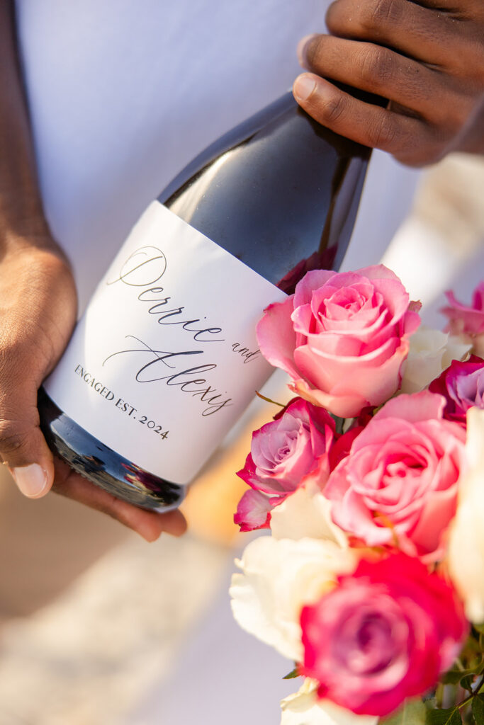 champagne bottle with a custom label and pink roses in a beach engagement photo at Jetty Park Beach in Cape Canaveral, Florida, captured by Snapshots by Gabriela