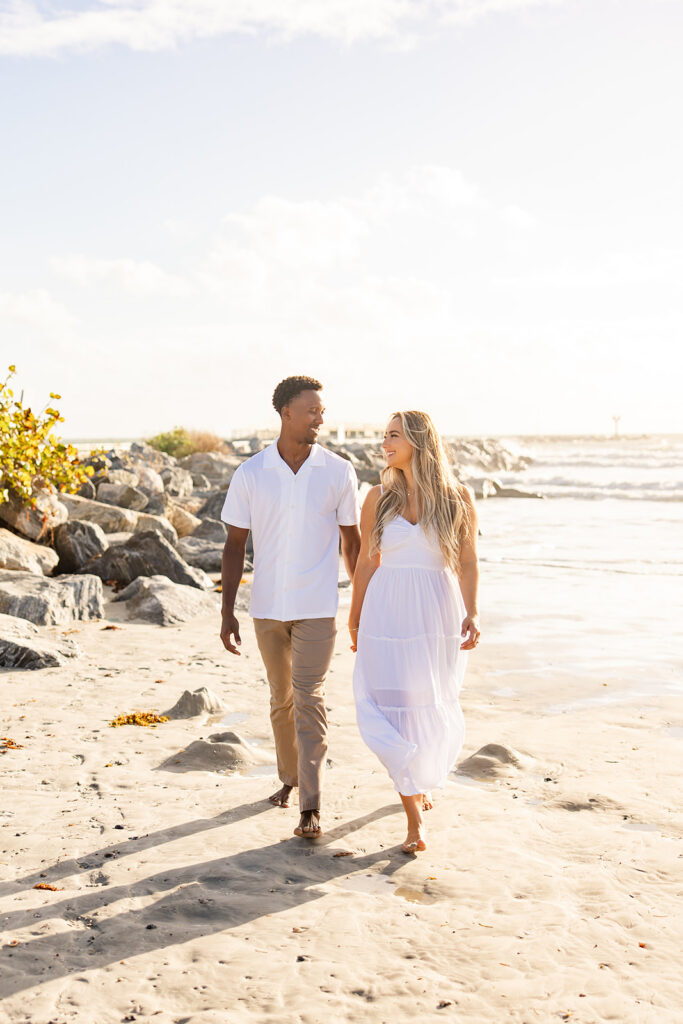 couple wearing white walk along the shore in a beach engagement photo at Jetty Park Beach in Cape Canaveral, Florida, captured by Snapshots by Gabriela