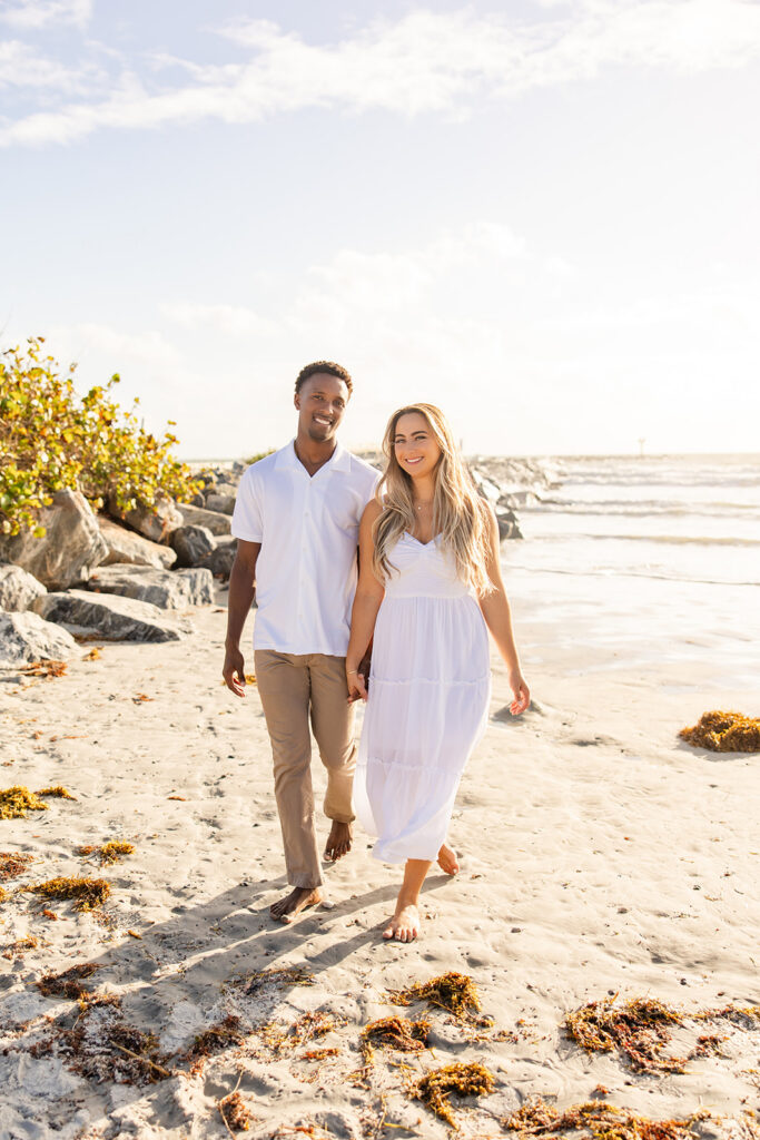 couple wearing white walk along the shore in a beach engagement photo at Jetty Park Beach in Cape Canaveral, Florida, captured by Snapshots by Gabriela