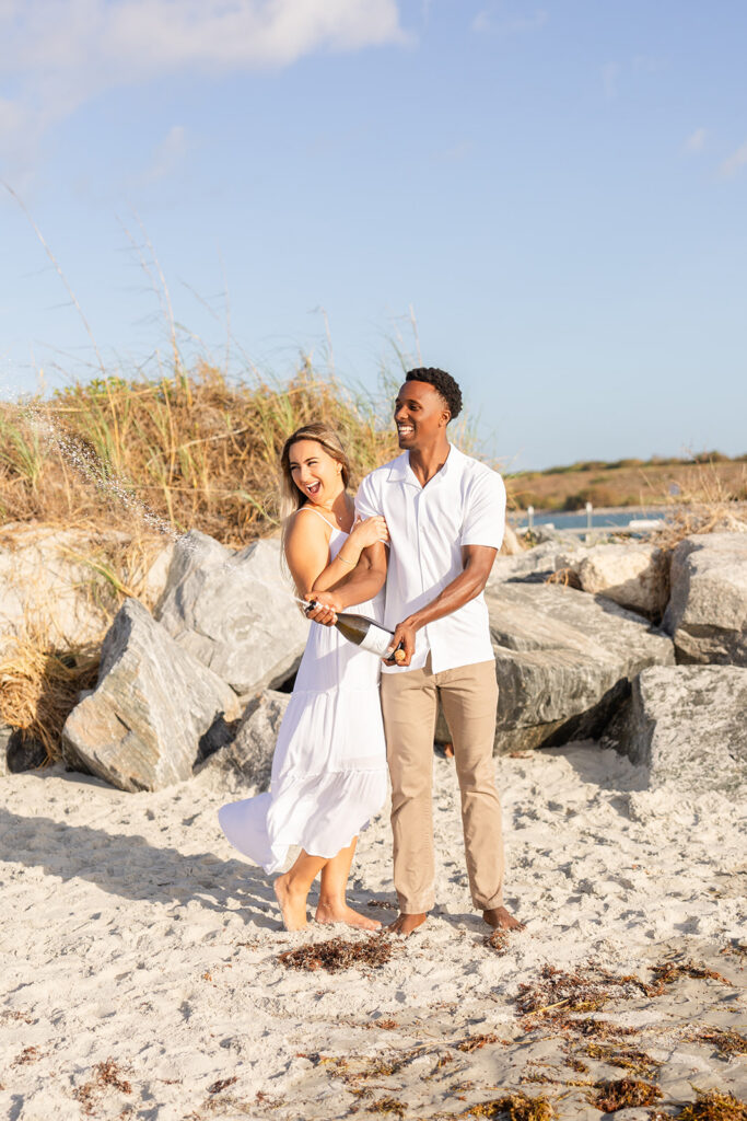 couple wearing white pop a champagne bottle in a beach engagement photo at Jetty Park Beach in Cape Canaveral, Florida, captured by Snapshots by Gabriela