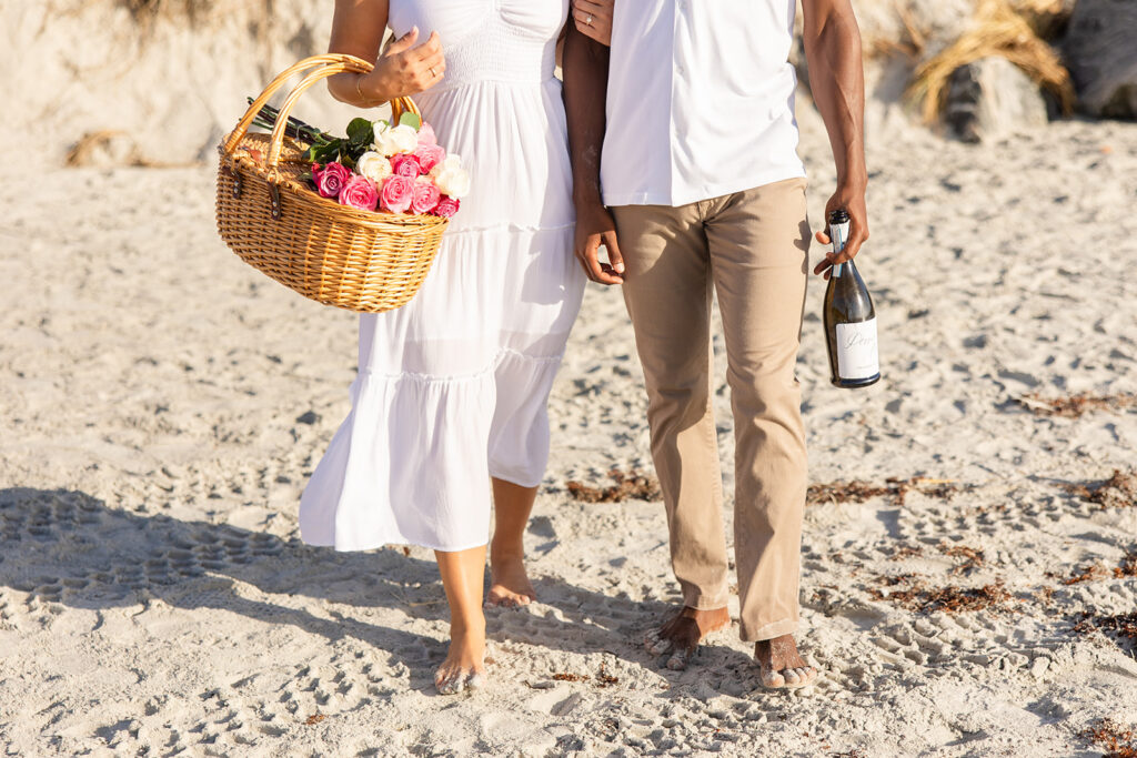 woman holding a picnic basket and man holding a champagne bottle walk along the shore in a beach engagement photo at Jetty Park Beach in Cape Canaveral, Florida, captured by Snapshots by Gabriela