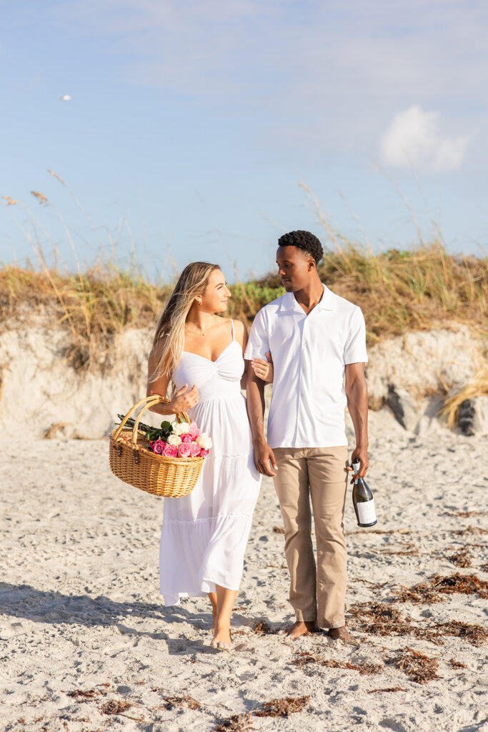 woman holding a picnic basket and man holding a champagne bottle walk along the shore in a beach engagement photo at Jetty Park Beach in Cape Canaveral, Florida, captured by Snapshots by Gabriela