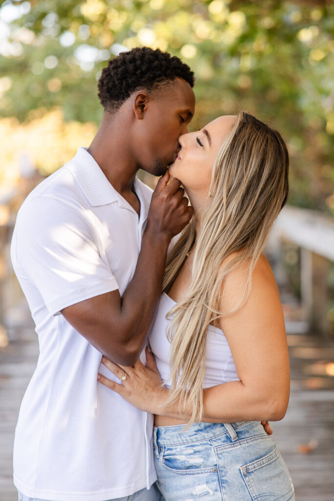 couple wearing white tops and jeans kiss on a wooden pier surrounded by greenery in a beach engagement photo at Jetty Park Beach in Cape Canaveral, Florida, captured by Snapshots by Gabriela
