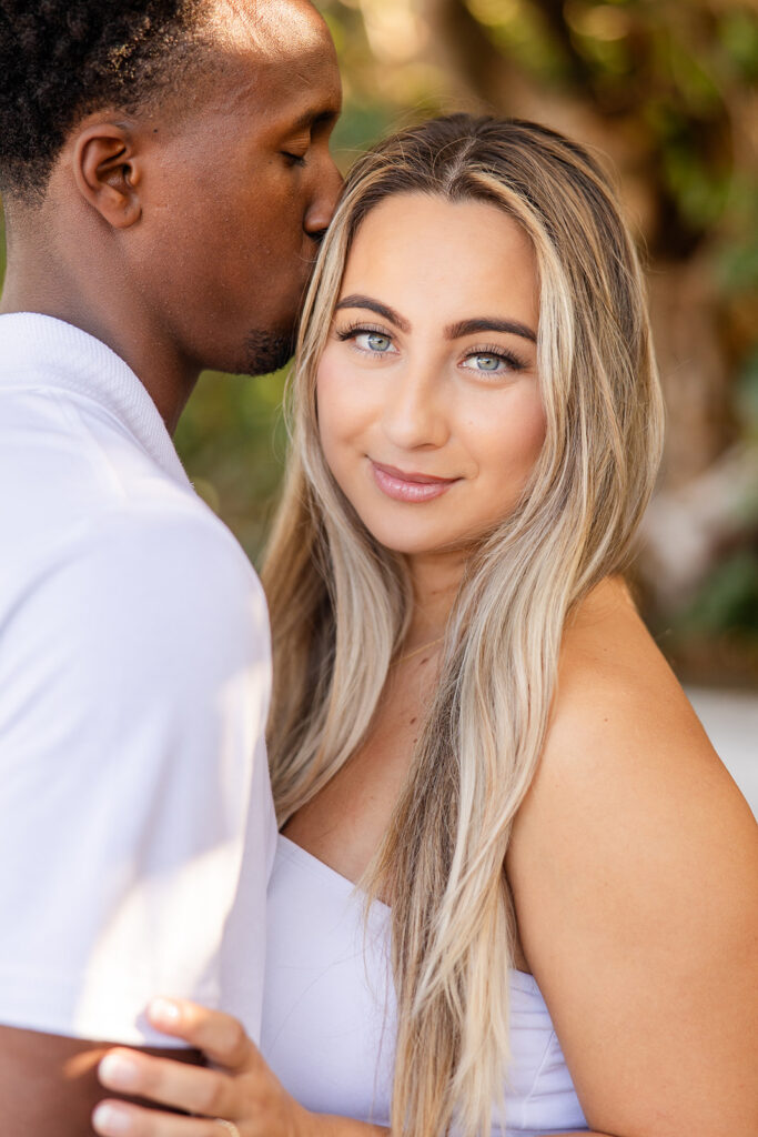 couple wearing white tops and jeans pose on a wooden pier surrounded by greenery in a beach engagement photo at Jetty Park Beach in Cape Canaveral, Florida, captured by Snapshots by Gabriela