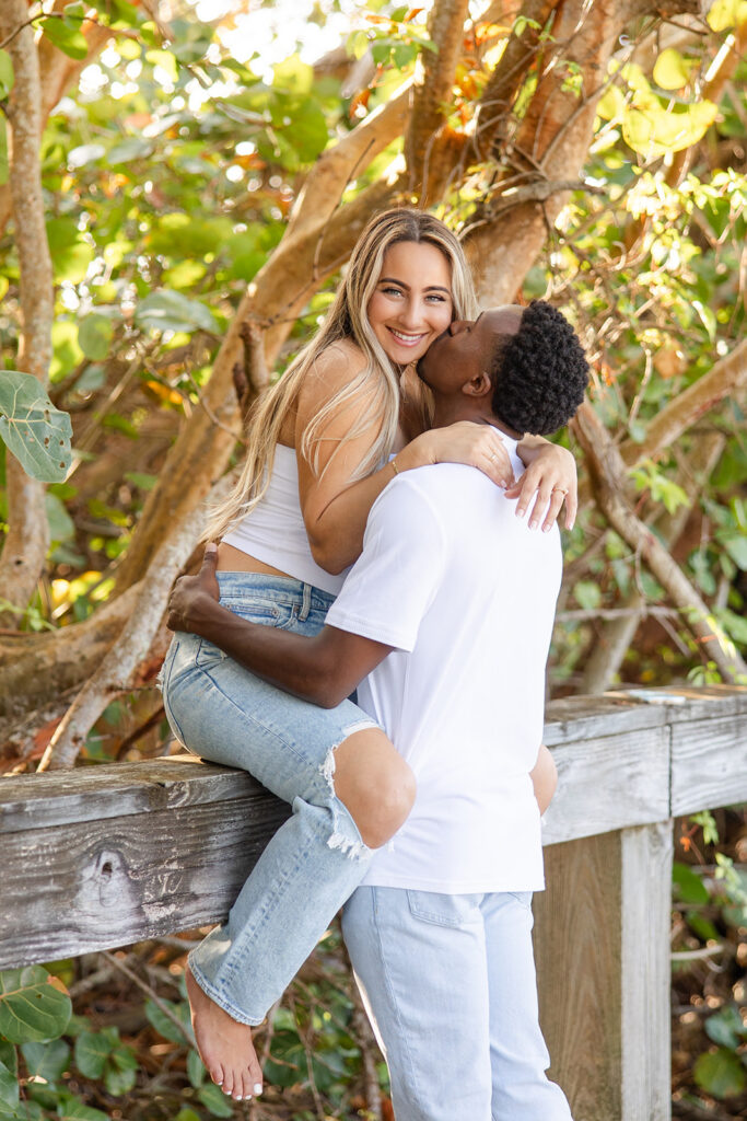 couple wearing white tops and jeans embrace on a wooden pier surrounded by greenery in a beach engagement photo at Jetty Park Beach in Cape Canaveral, Florida, captured by Snapshots by Gabriela
