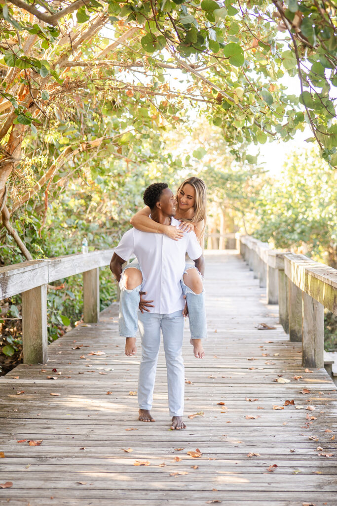 couple wearing white tops and jeans laugh while walking on a wooden pier surrounded by greenery in a beach engagement photo at Jetty Park Beach in Cape Canaveral, Florida, captured by Snapshots by Gabriela