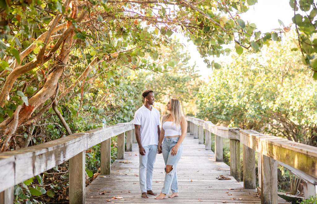 couple wearing white tops and jeans pose on a wooden pier surrounded by greenery in a beach engagement photo at Jetty Park Beach in Cape Canaveral, Florida, captured by Snapshots by Gabriela
