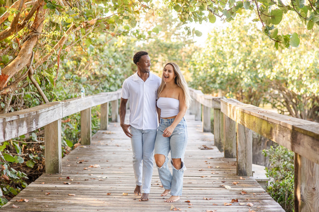 couple wearing white tops and jeans laugh while walking on a wooden pier surrounded by greenery in a beach engagement photo at Jetty Park Beach in Cape Canaveral, Florida, captured by Snapshots by Gabriela