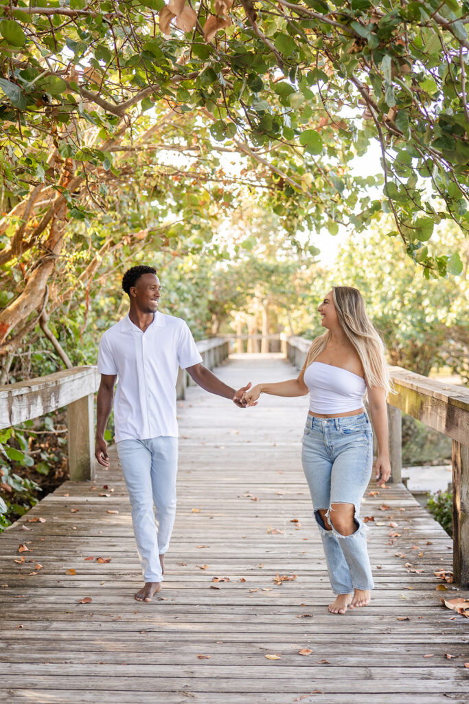 couple wearing white tops and jeans laugh while walking on a wooden pier surrounded by greenery in a beach engagement photo at Jetty Park Beach in Cape Canaveral, Florida, captured by Snapshots by Gabriela