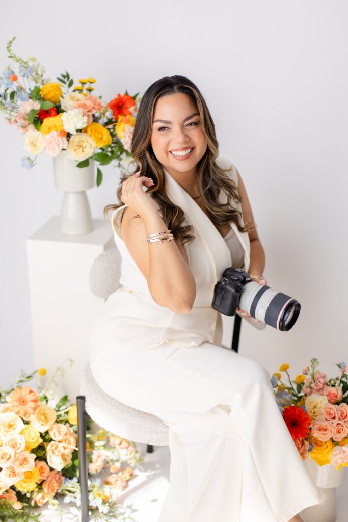a branding photo of Gabriela from Snapshots by Gabriela, an Orlando wedding photographer, posing while wearing white and holding her camera in front of colorful flowers