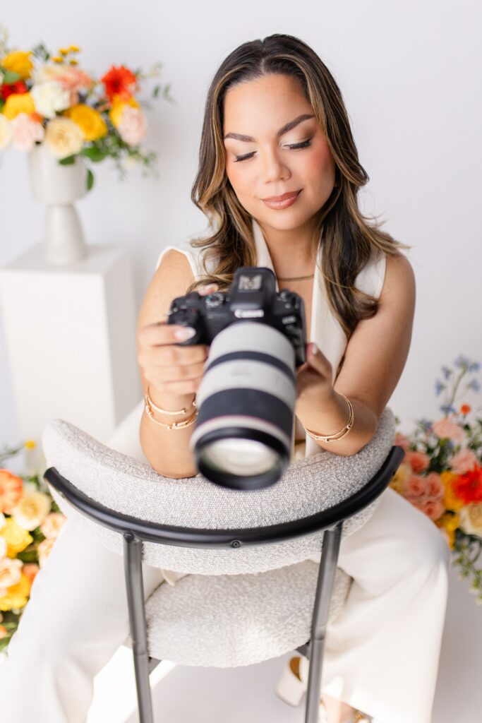 a branding photo of Gabriela from Snapshots by Gabriela, an Orlando wedding photographer, posing while wearing white and holding her camera in front of colorful flowers