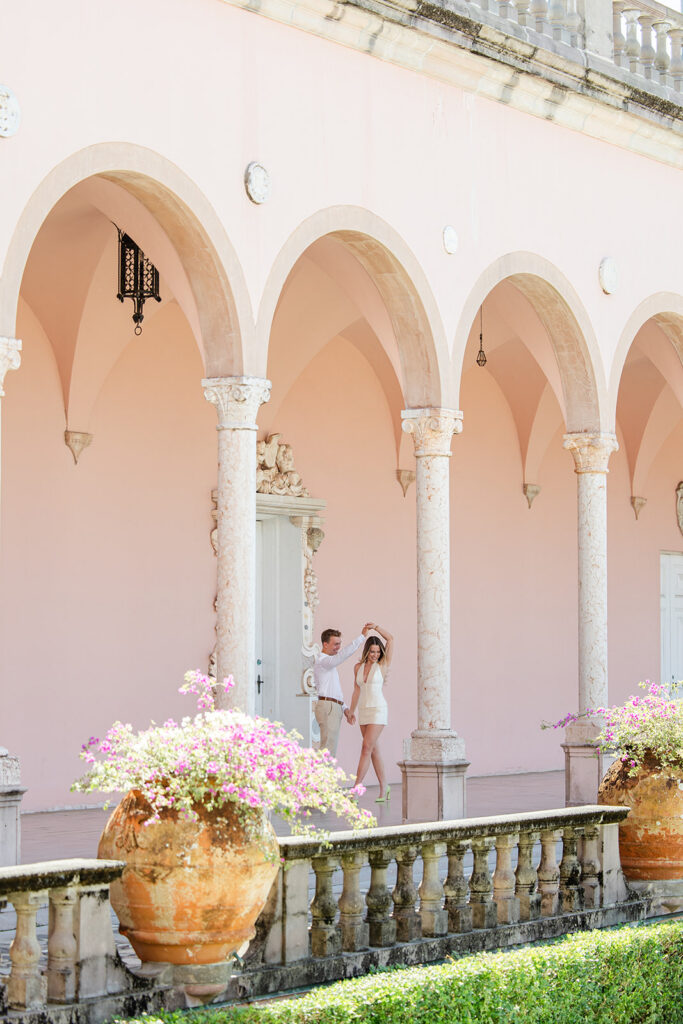 Woman in white mini-dress and man in white shirt and khaki pants under pink archways at The Ringling Museum in Sarasota for their engagement photos captured by Snapshots by Gabriela