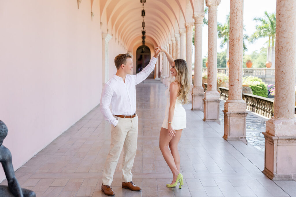 Woman in white mini-dress and man in white shirt and khaki pants under pink archways at The Ringling Museum in Sarasota for their engagement photos captured by Snapshots by Gabriela