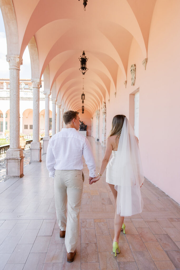 Woman in white mini-dress and bridal veil and man in white shirt and khaki pants kiss under pink archways at The Ringling Museum in Sarasota for their engagement photos captured by Snapshots by Gabriela