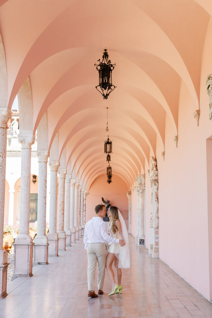 Woman in white mini-dress and bridal veil and man in white shirt and khaki pants kiss under pink archways at The Ringling Museum in Sarasota for their engagement photos captured by Snapshots by Gabriela