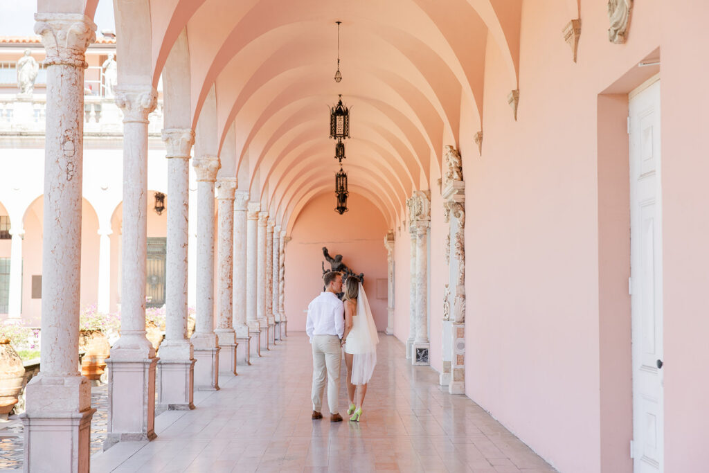 Woman in white mini-dress and bridal veil and man in white shirt and khaki pants kiss under pink archways at The Ringling Museum in Sarasota for their engagement photos captured by Snapshots by Gabriela